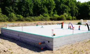Płyta fundamentowa koszt budowy domu. Betonowa konstrukcja z instalacjami hydraulicznymi i elektrycznymi, otoczona zieloną izolacją i ziemią.