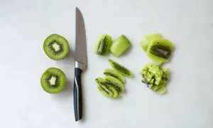Ein Messer liegt neben zwei halbierten Kiwis. Daneben sind die Früchte in Scheiben und Würfel geschnitten, bereit zum kiwi schneiden.