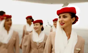 Uśmiechnięte stewardessy Emirates w czerwonych beretach i beżowych uniformach idą po płycie lotniska.