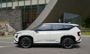 Nowy, biały suv Kia EV9 stoi zaparkowany przed nowoczesnym budynkiem z kamienną fasadą i dużymi oknami.