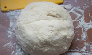 Surowe ciasto na pizzę na drewnianym blacie, posypane mąką. Obok żółta łopatka.