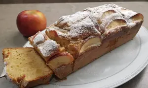Un delizioso plumcake alle mele, cosparso di zucchero a velo, con una mela rossa sullo sfondo.