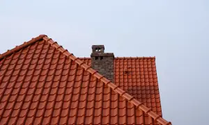 Ceglany komin, wyjście z pieca do komina, na tle jasnego nieba i pomarańczowego dachu.