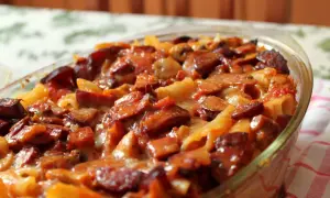 Pyszna zapiekanka makaronowa z kiełbasą, podana w szklanym naczyniu, kusi apetycznym wyglądem.