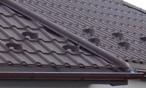 Brązowy dach z dachówki ceramicznej, widoczne elementy systemu rynnowego i wentylacyjnego. Jak zrobić zakończenie gąsiora?