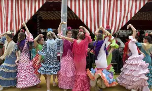 Niñas con trajes de volantes y lunares, aprendiendo a bailar sevillanas con arte bajo una carpa a rayas.