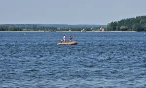 Dwóch wędkarzy na pontonie na szerokiej rzece na Roztoczu. W tle zielone wzgórza i zabudowania.