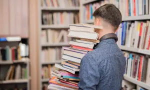 Młody mężczyzna w bibliotece dźwiga stos książek, symbolizując przejście od statusu ucznia do statusu studenta.