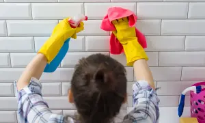 Ręce w żółtych rękawiczkach spryskują białe płytki i wycierają je różową ściereczką. Dowiedz się, jak usunąć uszczelniacz z płytek.