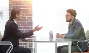 Dwóch mężczyzn w garniturach siedzi przy biurku. Jeden gestykuluje, jakby omawiał, czy na rozmowie kwalifikacyjnej pytać o zarobki.