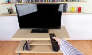 Ręka z pilotem sięga do telewizora naprzeciwko okna. Na półkach stoją płyty i kolorowe słoiki.