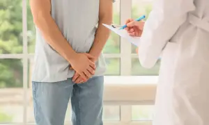 Mężczyzna w jasnej koszulce i jeansach stoi przed lekarzem w białym kitlu, który notuje coś na kartce. To wizyta kontrolna, być może związane z badaniem krwi na prostatę.