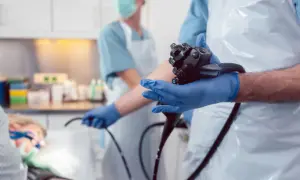 Lekarz w rękawiczkach przygotowuje się do cystoskopii, trzymając w dłoniach endoskop. W tle widać pacjenta i pielęgniarkę.