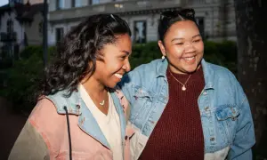 Twee vriendinnen lachen, maar de één voelt zich teleurgesteld in vriendschap. Ze dragen spijkerjacks en truien.