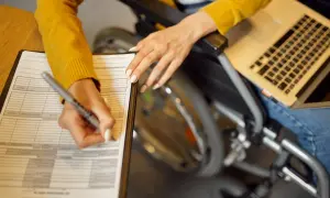 Kobieta na wózku inwalidzkim wypełnia dokumenty, korzystając z laptopa. To może być wniosek o 500 plus dla niepełnosprawnych.