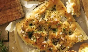 Pyszna focaccia z rozmarynem i solą morską, idealna do przygotowania według prostego przepisu. Obok kawałki pieczonych ziemniaków i plasterki wędliny.