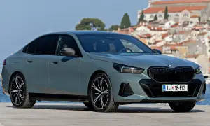 Szary sedan BMW 5 2025 z czarnym grillem i felgami, zaparkowany na tle malowniczego miasta nad morzem.
