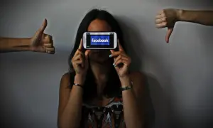 Una persona oculta su rostro tras un móvil con Facebook. A los lados, pulgares arriba y abajo, reflejando el impacto de las redes sociales y la autoestima.