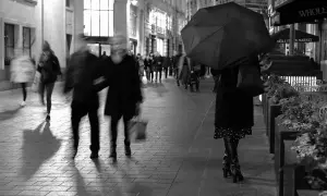 Nocna street photo. Rozmyte postacie spieszą się pod parasolem. W tle widać sklepy i światła miasta.