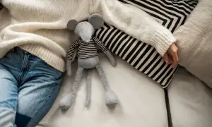 Graues Stofftier-Mäuschen mit gestreiftem Oberteil liegt neben Person in Strickpullover und Jeans auf Sofa.