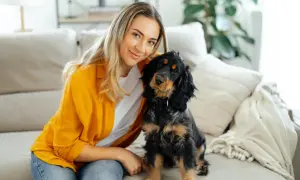 Uśmiechnięta blondynka i jej piesek siedzą na kanapie. Piesek patrzy w obiektyw, jakby chciał powiedzieć: "dlaczego pies chodzi za mną krok w krok?".