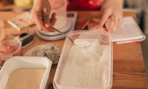 Ręce odmierzają mąkę do pojemnika, przygotowując ją do długoterminowego przechowywania. Na stole widać inne składniki i notatki.