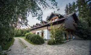 Urokliwa stajnia pod świerkami, otoczona zielenią, z kamienistą ścieżką prowadzącą do wejścia.
