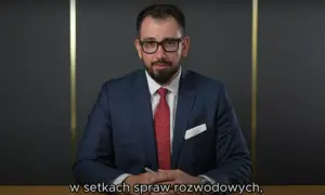Mężczyzna w garniturze i okularach siedzi przy biurku, trzymając długopis.