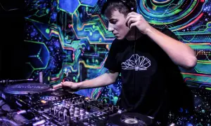 Młody DJ w słuchawkach miksuje muzykę na tle psychodelicznej, neonowej ściany.
