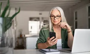 Frau mit Brille und Handy am Tisch, plant das Haus verkaufen und in Mietwohnung ziehen.