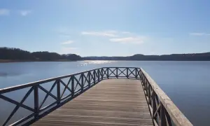 Holzsteg am Schmachter See lädt zum Spaziergang auf dem Rundweg ein. Sonnenschein glitzert auf dem Wasser, bewaldete Ufer im Hintergrund.