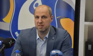 Dominik Kwapisiewicz w garniturze przed mikrofonami, na tle niebiesko-żółtego logo.