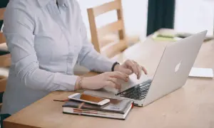 Kobieta w jasnej koszuli pracuje na laptopie, obok leży telefon i notatnik. Dba o swoje ubezpieczenie chorobowe.