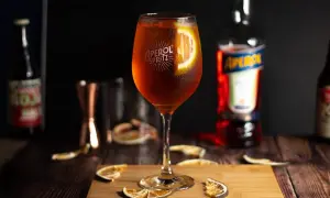 Orzeźwiający Aperol Spritz w kieliszku, ozdobiony plasterkiem pomarańczy. Obok butelka Aperol i suszone pomarańcze.