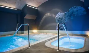 Piscinas de hidromasaje iluminadas con luz azul, con barandillas metálicas. Un mural de una escultura antigua adorna la pared, sugiriendo el mejor spa de Valencia.