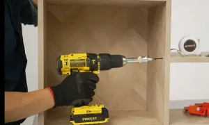Mano con guante negro usa taladro amarillo para unir dos muebles. Cinta métrica y herramientas al fondo.