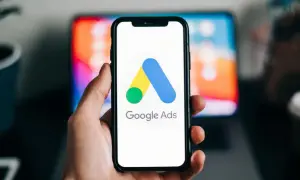 Dłoń trzyma smartfon z logo Google Ads. W tle widać ekran laptopa. Dziś budżet w reklamie Google Ads ustalamy.