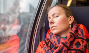 Kobieta w pomarańczowej kurtce i wzorzystym szaliku odpoczywa w pociągu, wyraźnie odczuwając zmęczenie.