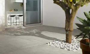 Un moderno patio con diversi tipi di pavimentazione esterna: lastre in gres grigio e un'area con ciottoli bianchi attorno a un albero.