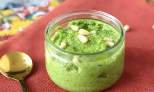 En burk med hemgjord egen pesto, toppad med pinjenötter. En guldig sked ligger bredvid på en röd duk.