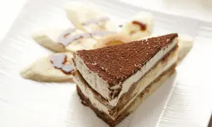 Tranche de tiramisu banane, saupoudrée de cacao, accompagnée de rondelles de banane nappées de sauce. Un délice !