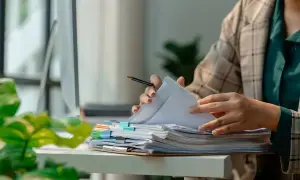 Pracownik przegląda dokumenty, marząc o urlopie dla poratowania zdrowia. Stos papierów czeka, a słońce wpada przez okno.