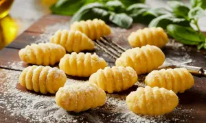Pyszne, domowe **makaron gnocchi** ułożone na drewnianej desce, posypane mąką. W tle zielona bazylia i widelec.