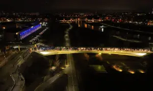 Nocne życie tętni na mostach berdychowskich. Tłumy ludzi spacerują po oświetlonym moście, a w tle widać miasto i rzekę.