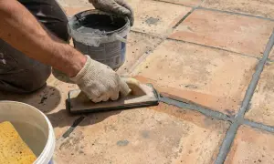 Application d'enduit pour joint carrelage extérieur. Un artisan, ganté, lisse le mortier gris avec une taloche sur des carreaux de terre cuite.