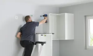 Hombre en escalera usa taladro para fijar soportes en pared, parte de como colocar kit para colgar armarios.