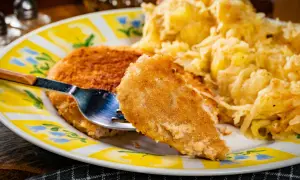Złocisty kotlet po parysku, chrupiący z wierzchu, z widelcem wbitym w kawałek. Obok porcja ziemniaków.
