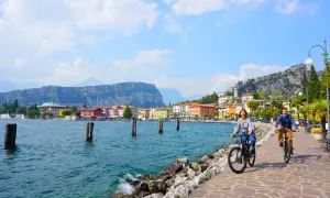 Coppia in bici sul lungolago di Riva del Garda, tra le attrazioni del posto, con case colorate e montagne sullo sfondo.