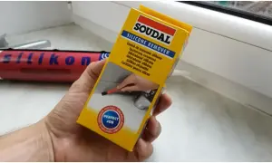 Ręka trzymająca opakowanie środka do usuwania silikonu Soudal. W tle widać tubkę z silikonem.
