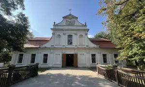 Biały kościół na wodzie w Zwierzyńcu, otoczony drzewami, z drewnianym mostkiem prowadzącym do wejścia.